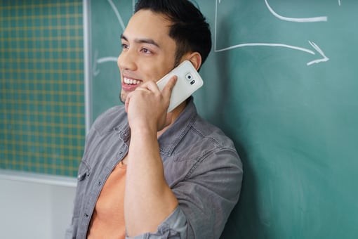 Young Asian man chatting on his mobile phone as he leans against a chalkboard in a school